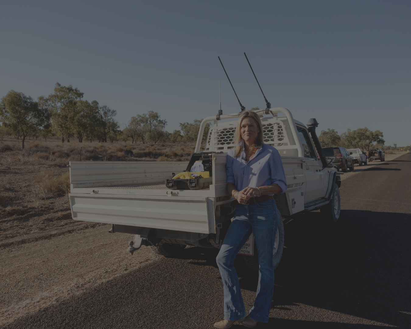 A woman in a blue shirt and jeans stands beside a white utility pickup truck parked on a rural road, with trees and another vehicle in the background under a clear sky.
